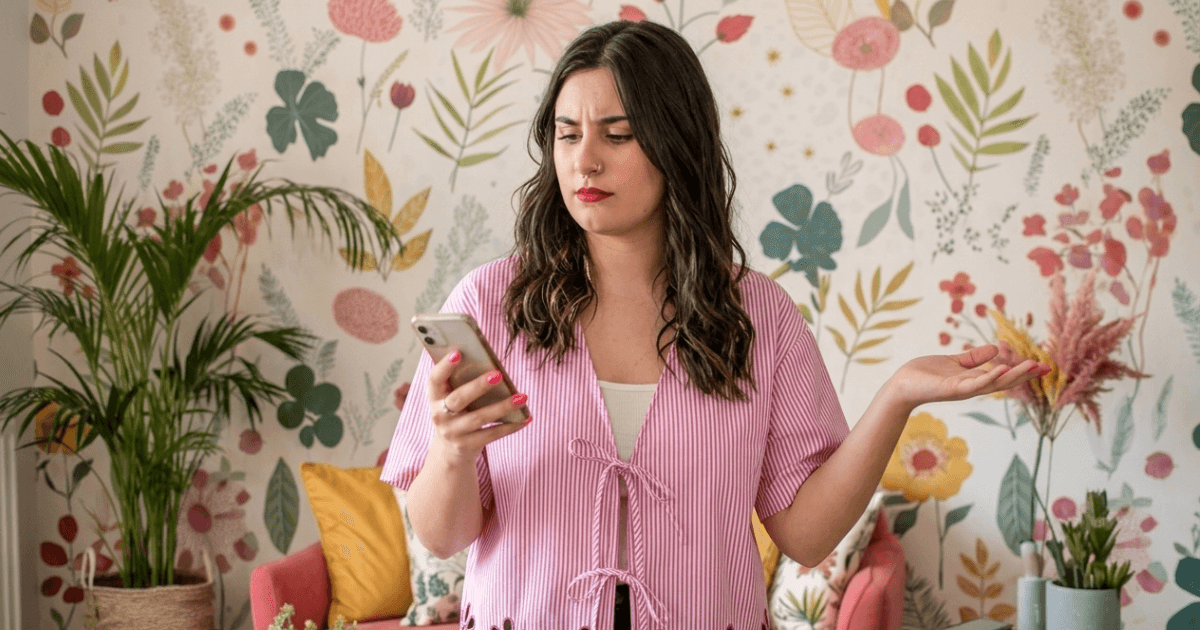 Mujer con blusa de rayas sostiene un teléfono móvil y muestra una expresión de desconcierto, en un ambiente colorido con plantas y un fondo de flores, reflejando la búsqueda de claridad en crear conte