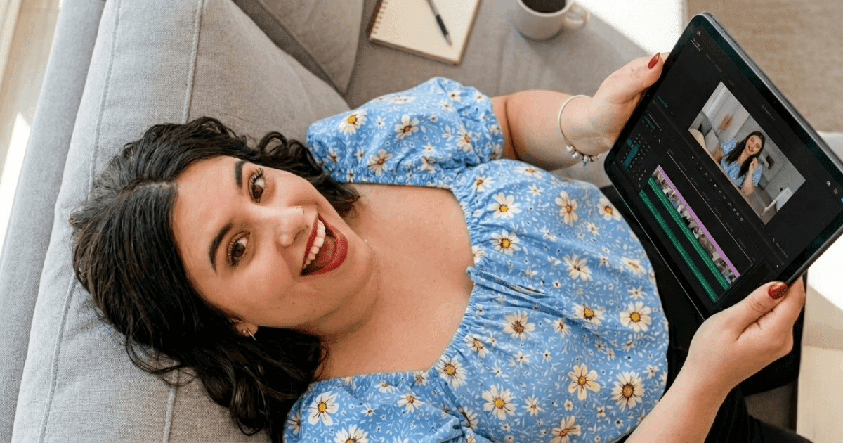Mujer sonriente con blusa azul con flores sostiene una tablet mostrando un programa de edición de video, sentada en un sofá, reflejando el uso de herramientas de IA para crear contenido para redes soc